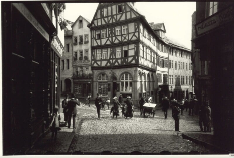 Black and white photo of a cobblestone street in a European town, with people walking, children playing, and a half-timbered building in the background. Other buildings and shop signs line the street.