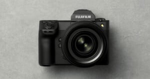 A black Fujifilm digital camera with a large lens is placed on a gray textured surface, viewed from above.