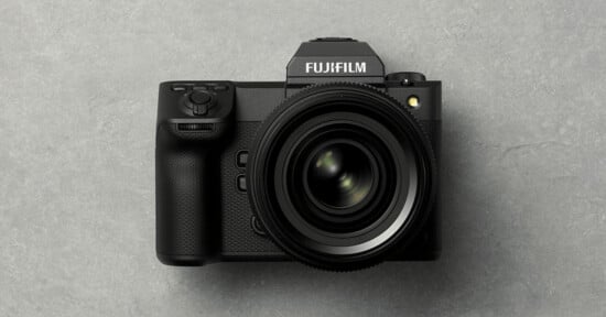 A black Fujifilm digital camera with a large lens is placed on a gray textured surface, viewed from above.