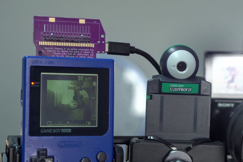 A blue Game Boy Pocket with a circuit board attached displays a pixelated camera view on its screen. Next to it is a Game Boy Camera accessory mounted on a camera tripod.