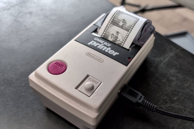 A close-up of a Nintendo Game Boy Printer with a photo strip being printed. The device is beige with a pink "Feed" button, a power switch, and a partially printed image visible on the paper.