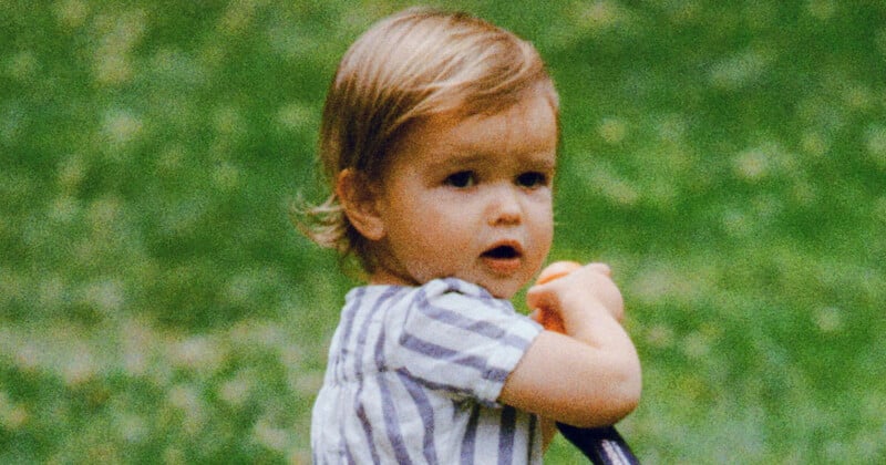 A young child with light brown hair, wearing a short-sleeved striped shirt, looks to the side while holding onto a black object, with a blurred green outdoor background.