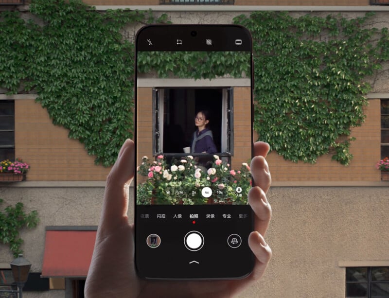 A hand holds a smartphone, capturing a photo of a woman smiling at a window surrounded by flowers. The building behind the window is covered with green ivy and has a brick exterior.