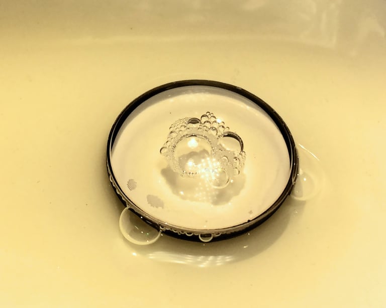 A metallic sink drain stopper with soap bubbles on its surface sits in a shallow pool of water, reflecting light in a yellowish sink basin.