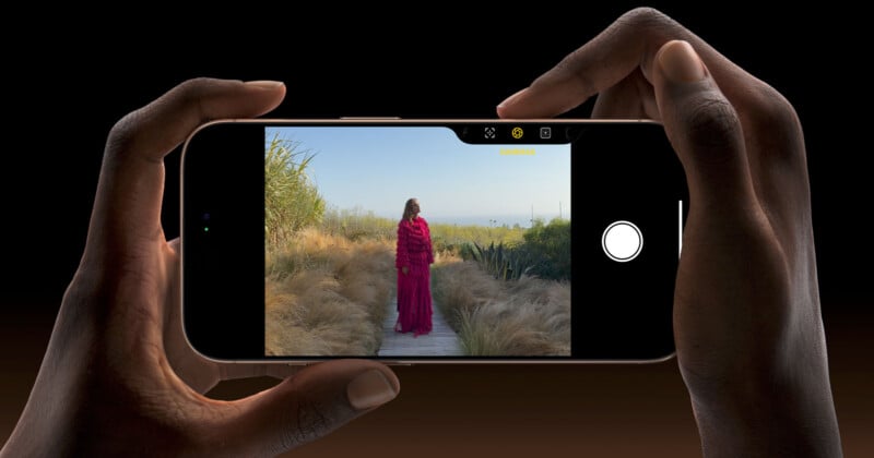 A person holds a smartphone horizontally, taking a photo of someone in a bright red outfit standing on a wooden path surrounded by tall grass and plants under a clear sky.