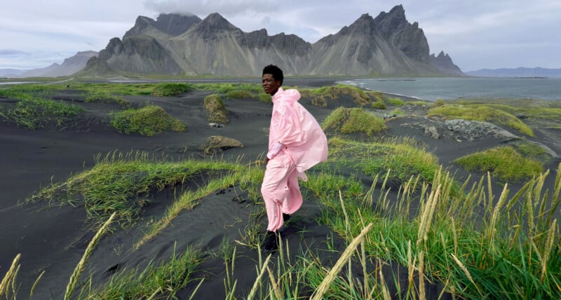 A person in a pink outfit stands on black sand with green grass, against a dramatic backdrop of jagged mountains and cloudy sky near the coast.