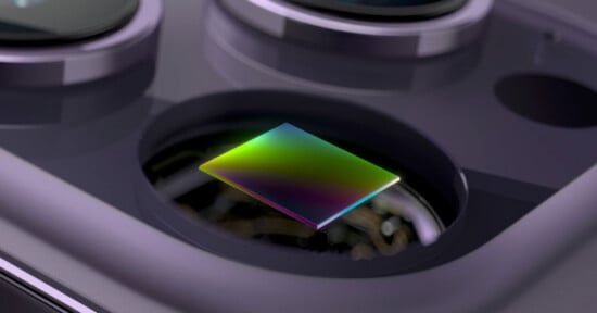 A close-up of a smartphone camera module showing a colorful, reflective sensor beneath the camera lens. The surface of the sensor displays a gradient of green, blue, and purple hues.