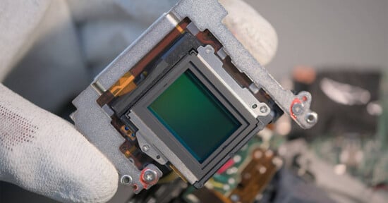 A gloved hand holds a digital camera image sensor surrounded by a metallic frame, showing intricate electronic components and circuitry.