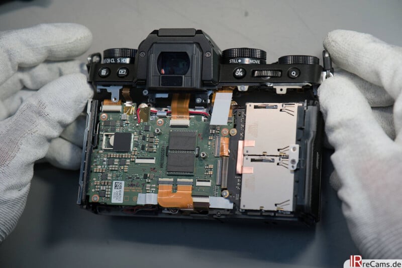 A pair of gloved hands holds an open digital camera, revealing its internal circuit board, cables, and dual SD card slots on a gray surface.