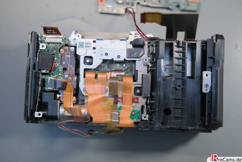 An opened electronic device, likely a camera, showing internal components, circuit boards, wires, and ribbon cables. The outer casing is partially removed, revealing the intricate internal structure.