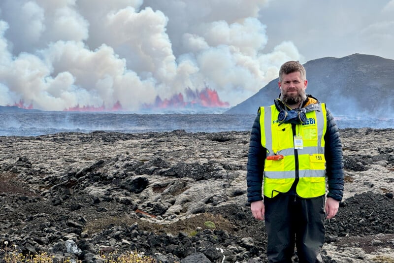 A man in a yellow safety vest stands on rocky ground with a volcanic eruption and billowing smoke in the background.