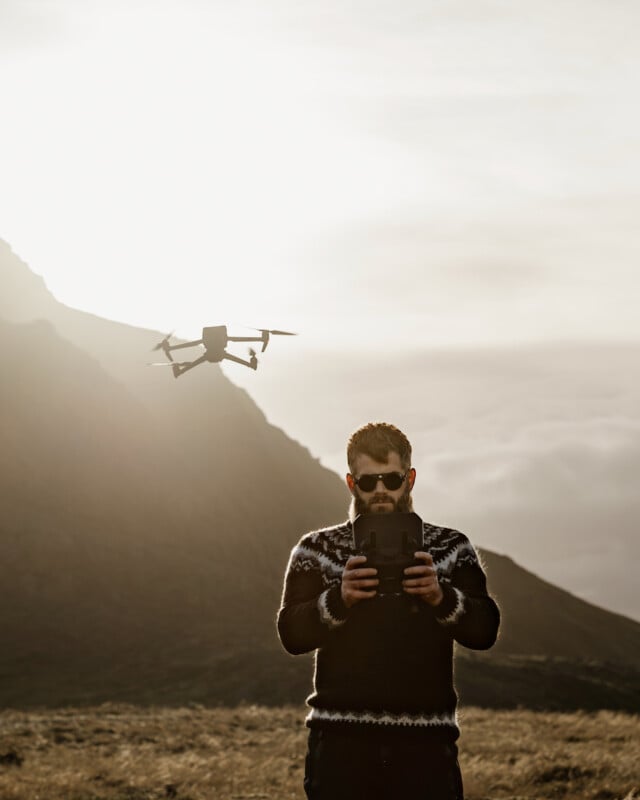 A man wearing sunglasses and a patterned sweater operates a drone with a remote controller in a mountainous landscape at sunset. The drone hovers in the air nearby.