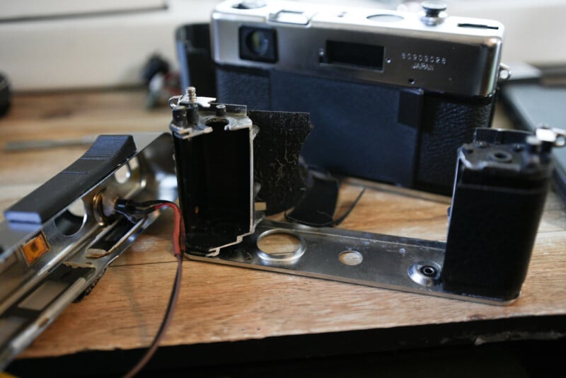 A partially disassembled film camera sits on a wooden surface, with its metal components and internal wiring exposed. Another intact camera is visible in the background.