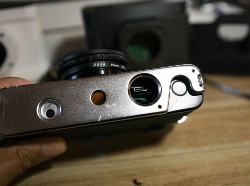Close-up of the bottom of a camera showing a USB-C charging port, a round battery compartment, a tripod mount, and part of the lens. The camera is being held above a wooden surface.