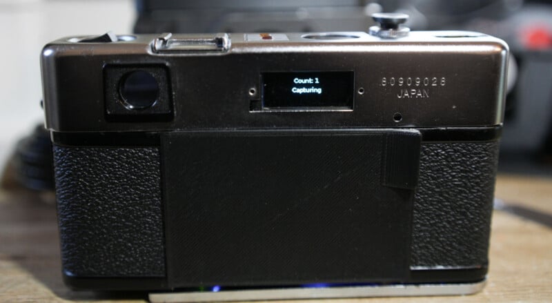 A close-up of the back of a vintage film camera with a digital screen displaying "Count: 1 Capturing." The camera is black with silver accents and is resting on a surface.