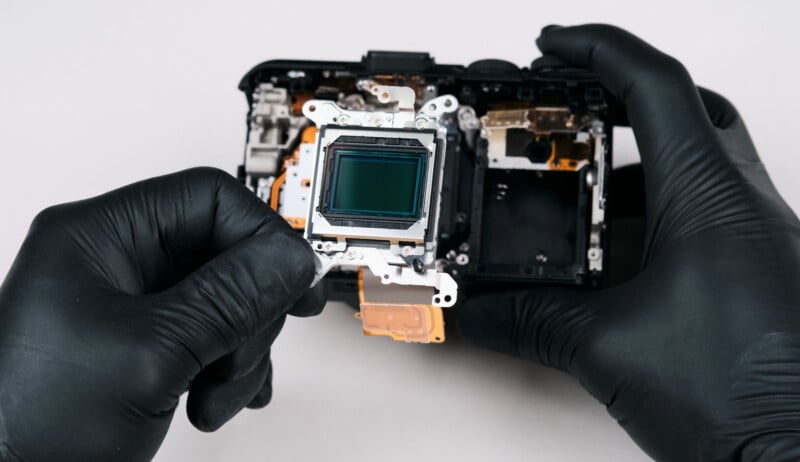 A person wearing black gloves holds a digital camera sensor, with the partially disassembled camera body visible in the background on a white surface.