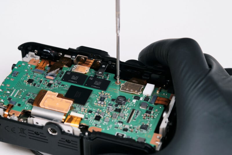 A person wearing a black glove uses a screwdriver to repair or assemble the green circuit board inside an electronic device.