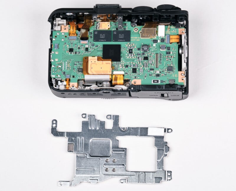A digital camera with its back cover removed, exposing the internal green circuit board and components, with a detached metal shield placed below it on a white surface.