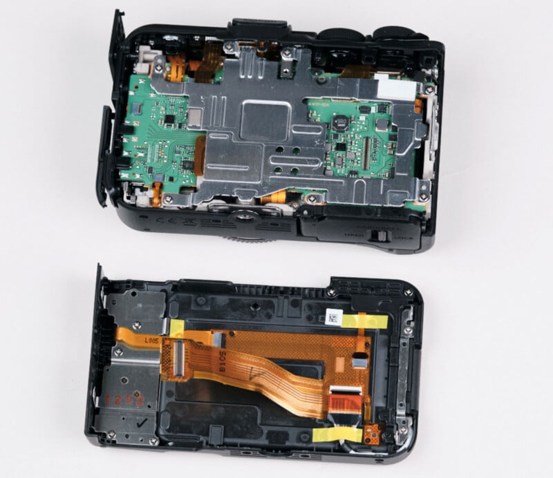 An open digital camera with its back cover removed, revealing internal circuit boards, wiring, and components. The back cover, with attached ribbon cables, is placed next to the camera body.