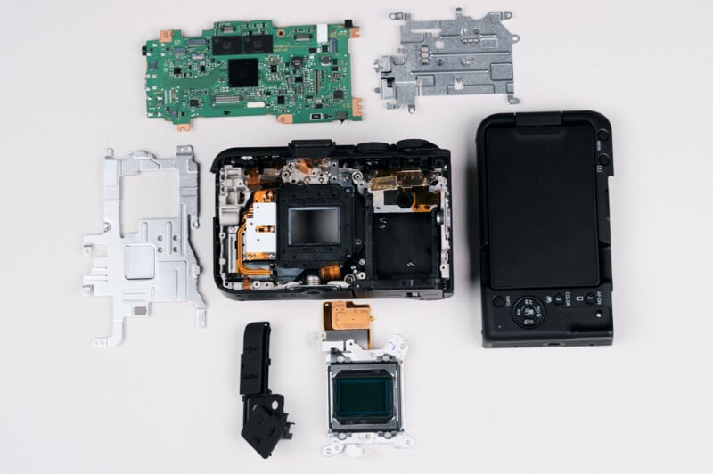 A digital camera is disassembled on a white surface, displaying its circuit boards, metal frames, sensor, and external casing arranged neatly around the opened main body.