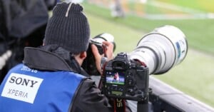 A sports photographer wearing a beanie and Sony XPERIA vest takes photos with a camera. Another camera in front shows an image of athletes on its display, with a football field visible in the background.