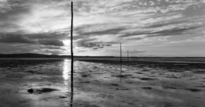 Black and white photo of a tidal flat at sunset, with tall wooden poles receding into the distance, reflections on wet sand, and dramatic clouds in the sky.