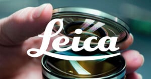 A hand holds a camera lens with reflections visible on the glass. The white Leica logo is prominently overlaid across the center of the image.