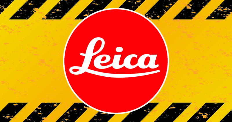 A red oval with the white Leica logo in the center, set against a yellow background with black diagonal hazard stripes along the top and bottom edges.