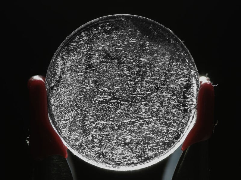 A pair of gloved hands holds a circular, transparent disc with many scratches and lines, illuminated from behind against a dark background.