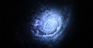 A swirling spiral of bright, bluish-white clouds or gas illuminated against a dark, almost black background, resembling a vortex or galaxy in space.