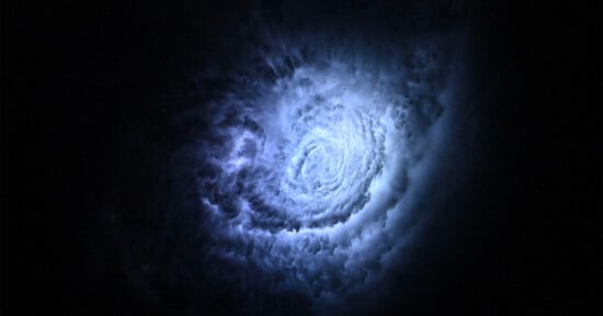 A swirling spiral of bright, bluish-white clouds or gas illuminated against a dark, almost black background, resembling a vortex or galaxy in space.