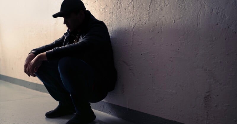 A person wearing a cap and jacket sits crouched against a wall in a dimly lit, empty corridor, appearing deep in thought or distressed, with their face mostly in shadow.