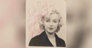 Black and white portrait of a woman with short, wavy blonde hair and a dark top, featuring a handwritten red autograph across the top left side of the photo.