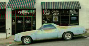 A rusty, light blue vintage car is parked in front of a bike and skate shop with striped awnings and large storefront windows displaying white painted signs.
