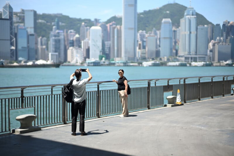 Two people pose for a photo by a waterfront railing, with a city skyline and skyscrapers in the background across the water on a sunny day.