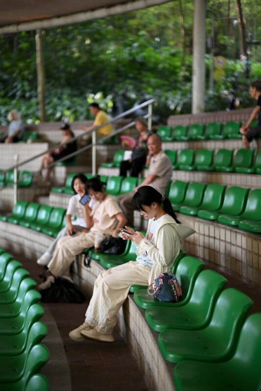 Several people sit scattered on green plastic seats in an outdoor amphitheater. Three young women sit together, two talking and one using her phone, while others are seated further back. Lush greenery is visible in the background.
