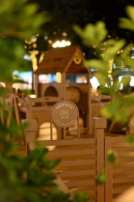 A softly lit outdoor playground with a wooden playhouse in the background and a sign reading "Pet Cart Parking" in English and Chinese in the foreground, partially framed by leafy green plants.