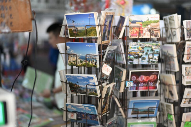 A display rack filled with colorful postcards featuring various cityscapes and landmarks stands at a market stall, with a blurred person and more cards visible in the background.