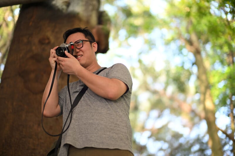 A man wearing glasses and a gray shirt smiles while taking a photo with a camera outdoors, with sunlight filtering through green trees in the background.