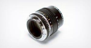 A close-up photo of a camera lens with detailed focus and aperture markings, placed on a white background. The lens is angled to show both the glass and the metallic mount.