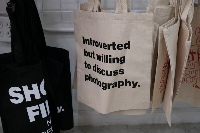 A beige tote bag hangs on display with the text "Introverted but willing to discuss photography." Other tote bags with partial text are visible beside it.