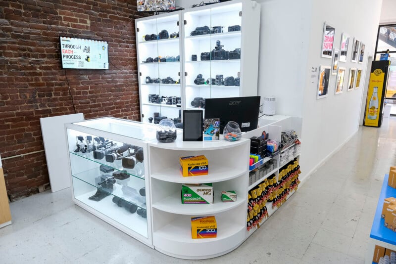 A camera store counter with glass display cases holding cameras and lenses, shelves with film boxes, a computer monitor, and camera accessories. Behind, a tall shelf displays more cameras against a brick wall.