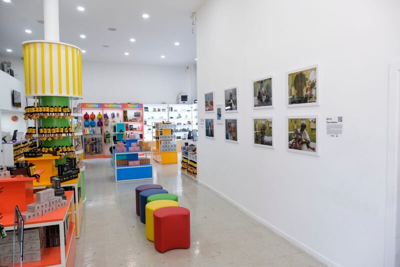 A brightly lit store interior with colorful shelves, toys, and gift items on display. Four cube-shaped stools are in the center, and framed photos hang on the white wall to the right. The decor is playful and modern.