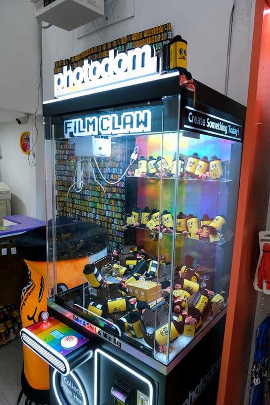 A claw machine labeled "photoodom FILM CLAW" is filled with various rolls of camera film instead of traditional prizes. The machine is brightly lit and sits in a modern, colorful shop.