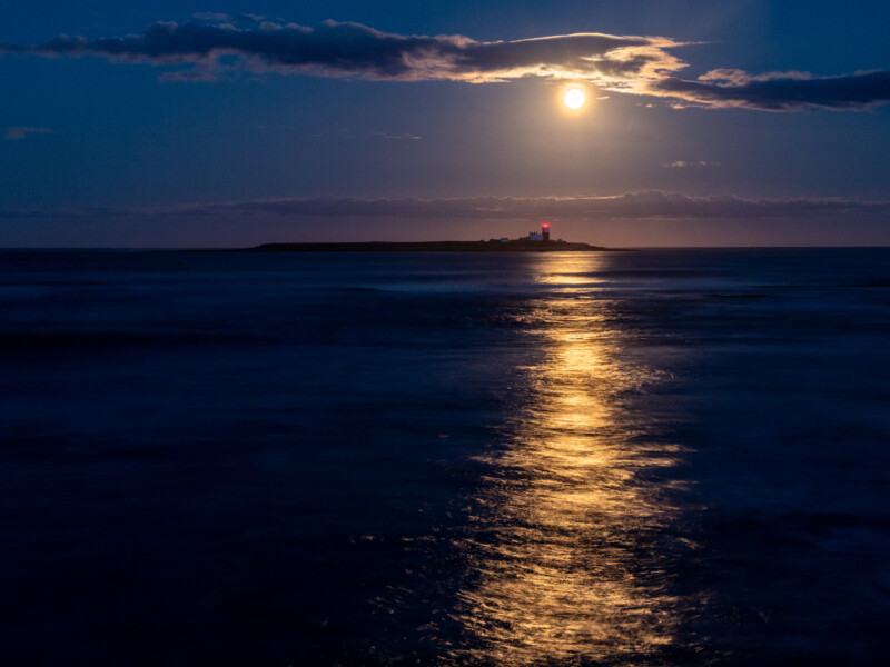 Full moon shines over the sea, casting a bright reflection on the water. In the distance, an island with a lighthouse is illuminated against the dark horizon under a cloudy night sky.