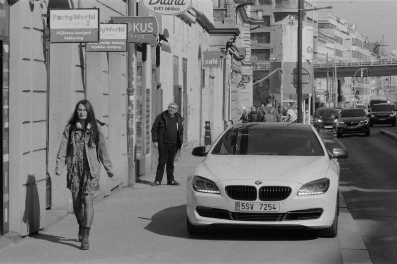 A woman walks on a city sidewalk past a white BMW car parked at the curb. Several people are further down the street, and buildings with signs in a foreign language line the road. The scene is in black and white.