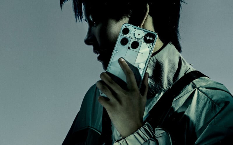 A person in a light jacket holds a smartphone with a unique, transparent back design and visible components, featuring circular camera modules, up to their ear. The image is dark and moody, with dramatic lighting.