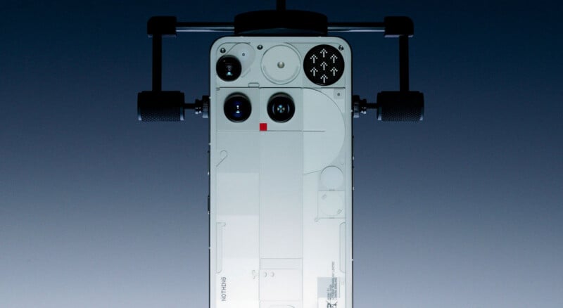 A white smartphone, viewed from the back, is attached to a horizontal camera rig with handles on both sides, against a gradient dark background.