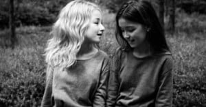 Two young girls sit closely together outdoors, smiling and looking at each other. One has light wavy hair and the other has straight dark hair. Both wear long-sleeved tops. The image is in black and white.