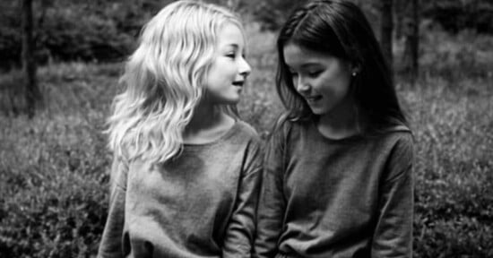 Two young girls sit closely together outdoors, smiling and looking at each other. One has light wavy hair and the other has straight dark hair. Both wear long-sleeved tops. The image is in black and white.